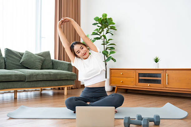 istock Young Asian woman stretching yoga workout on exercise mat while online training class with computer laptop in living room 2220506692