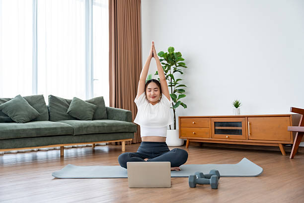 istock Young Asian woman stretching yoga workout on exercise mat while online training class with computer laptop in living room 2220506759
