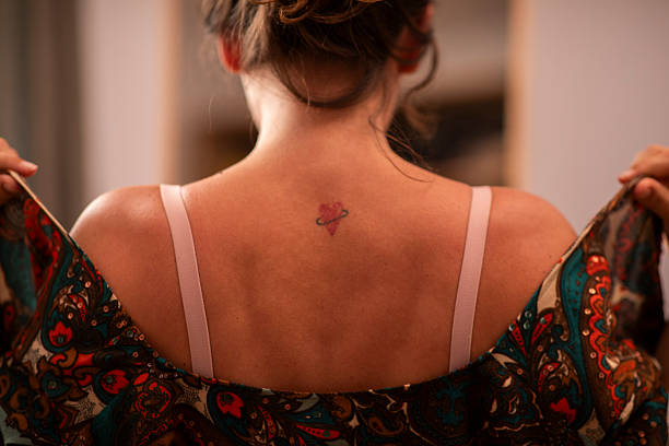 A woman with her back turned, featuring a delicate tattoo on her upper back, adjusts a colorful patterned blouse