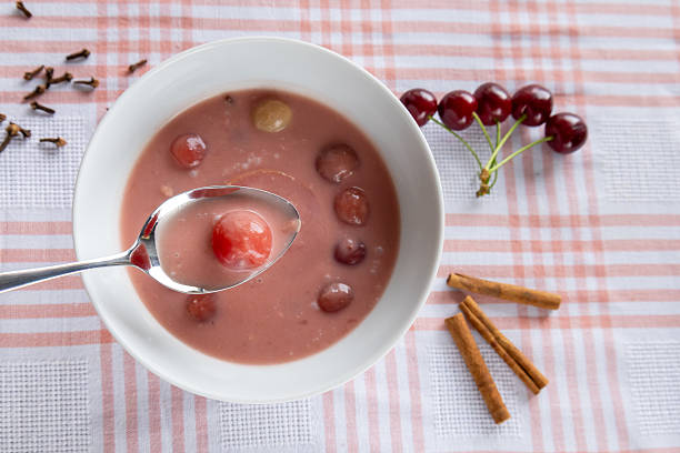 Overhead view of a bowl of chilled cherry soup served with whole fruits, with a spoon holding one bright cherry. The scene is enhanced by scattered cloves, cinnamon sticks, and fresh cherries on a red-and-white checkered cloth, evoking a homemade, summery feel. A traditional Hungarian cherry soup, easily adaptable for vegetarian and vegan diets.