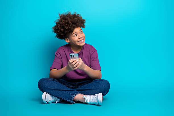 istock Young boy sitting cross legged holding smartphone smiling on blue background showing happiness and leisure 2220535797