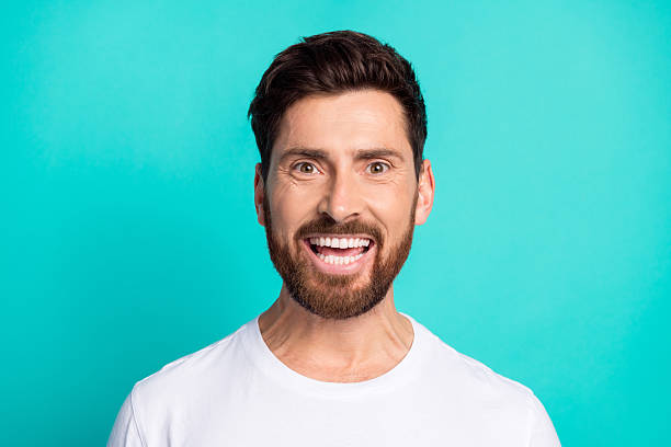 istock Handsome young man with a beard smiling brightly, dressed casually in a white shirt, standing against a teal background. 2220540280