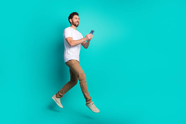 istock Young bearded man holding a smartphone, walking confidently against a turquoise background, showcasing modern lifestyle 2220540389