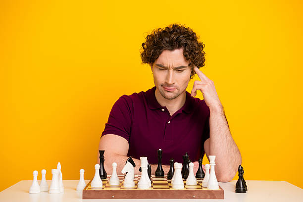 istock Young man thoughtfully playing chess on a bright yellow background, showcasing concentration and strategy in casual attire 2220540511
