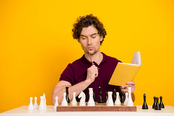 istock Young man reading book while analyzing chess moves on chessboard against vibrant yellow background 2220540514