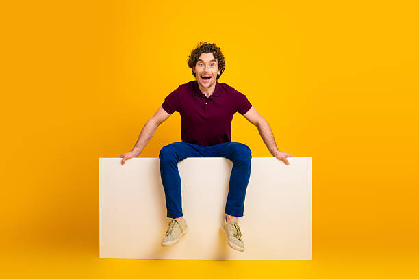 istock Engaged young man sitting on a white panel in front of yellow background conveying joyful energy and positivity 2220540519
