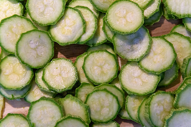 A high-angle close-up showcases neatly arranged zucchini slices on a rustic wooden surface. The uniform cuts reveal the fresh, light green flesh and slightly darker rind of the vegetable. The soft, even lighting highlights the vibrant colors and textures, suggesting food preparation or healthy eating.