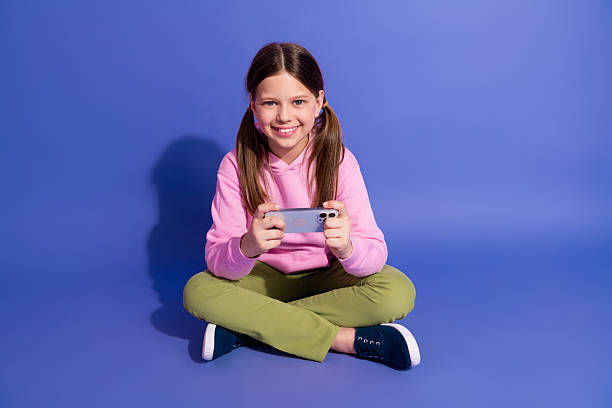 Smiling young girl using a smartphone, dressed in a casual pink hoodie and green pants, sitting on a purple background