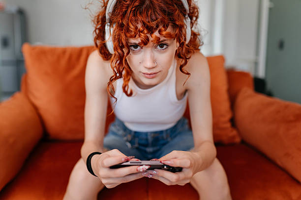 Young woman using mobile phone at home, sitting on couch and playing video games