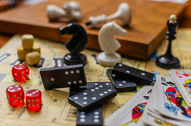 A close-up still life of various classic board games. The scene features a collection of leisure and hobby items including dominoes, red dice, playing cards and chess pieces.