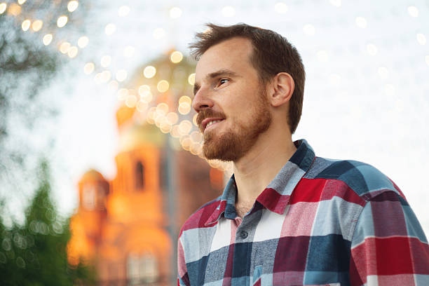 Smiling man enjoying festive evening lights in city. Guy with beard and modern hairstyle wearing in blue, red and white plaid shirt walking through the city streets in urban background.