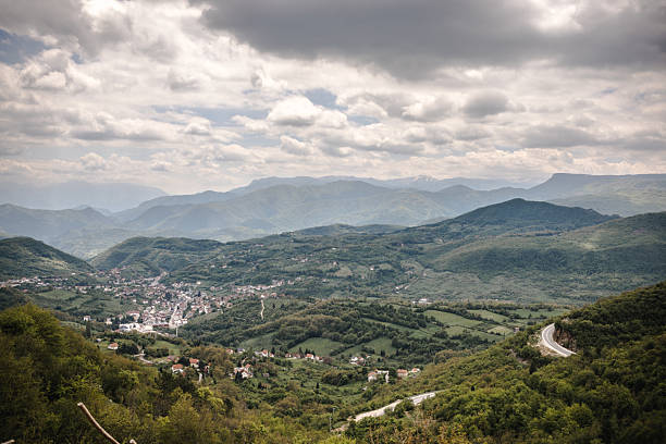 Picturesque view of a mountain valley, featuring a small settlement surrounded by expansive lush greenery, and rolling hills, captured under an overcast sky.