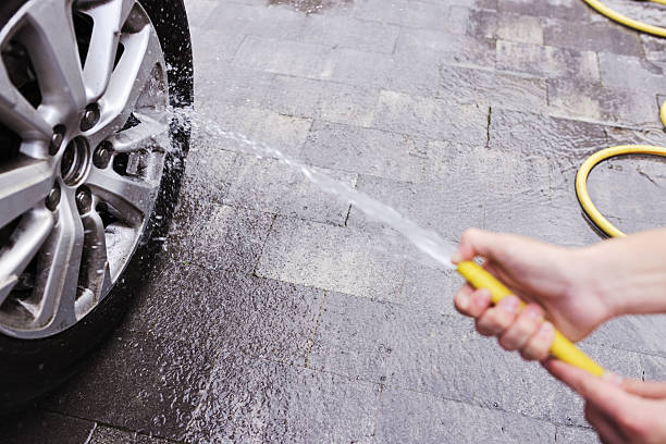 A person uses a garden hose to rinse a car wheel, cleaning it thoroughly while water splashes. The scene is set on a paved driveway, showcasing the process of vehicle maintenance at home.