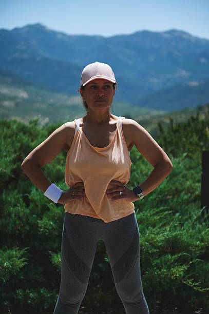 A sportily dressed woman stands confidently outdoors with hands on hips, backed by a stunning mountain view. She wears activewear and demonstrates energy and determination in a natural setting.