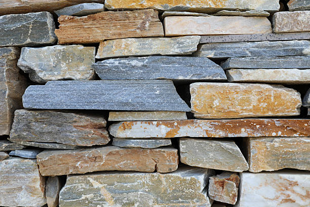 Close-up view of a dry stone wall constructed from irregularly shaped flat stones. The stones vary in color from gray and beige to rusty orange and off-white, showcasing natural textures and rough surfaces. Carefully stacked without visible mortar, the wall has a rustic, sturdy appearance, typical of traditional stone masonry.