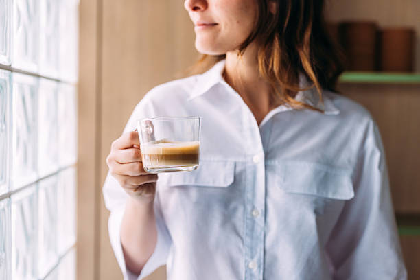 A woman wearing a white shirt holds a glass of coffee. The room is softly lit by sunlight, creating a warm and calm atmosphere. Her peaceful expression suggests relaxation.