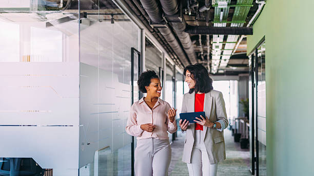 Two professional women walking in an open modern office environment, conversing and smiling. They appear engaged and positive, highlighting teamwork, professionalism, and a collaborative office culture.