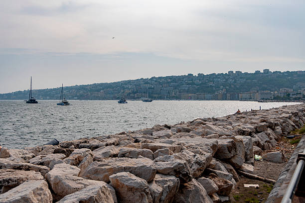 People enjoy a warm summer day on the seafront of Naples, Italy. The beach and rocks are full of locals and tourists relaxing and swimming, with the cityscape rising in the background