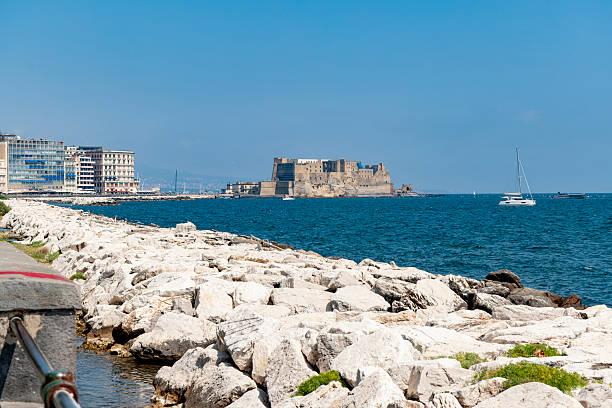 A view of Castel dell’Ovo from the Naples seafront, where the rocky shore meets the deep blue waters of the Gulf of Naples on a sunny day