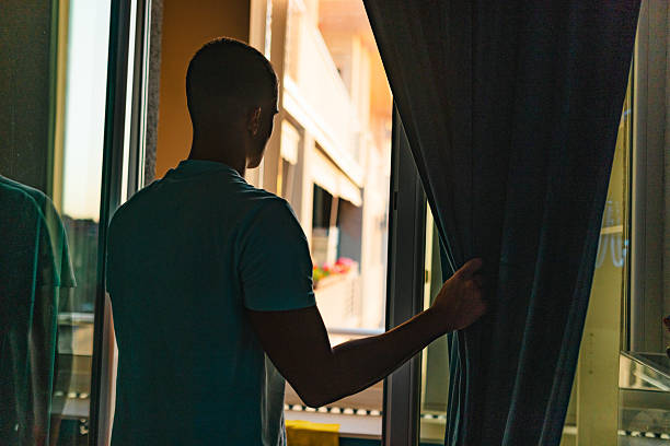 A person stands near a window, gently closing a curtain, with sunlight streaming in from the outside, illustrating a serene and thoughtful moment.