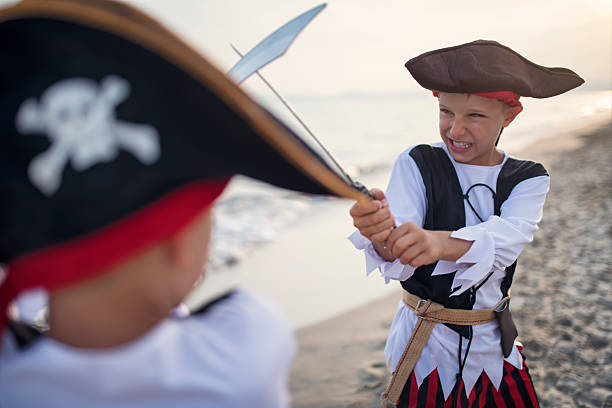 Two children are immersed in an imaginative sword fight on a sandy beach, fully embracing their pirate personas. Both children wear pirate hats gripping toy swords with playful determination.\nNikon D850