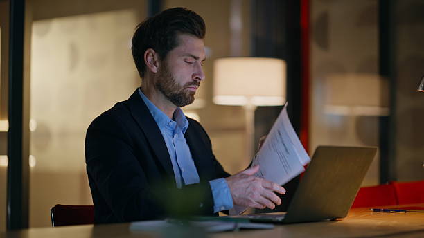 istock Late worker reading papers working at laptop in dark office closeup. Serious man 2220611558