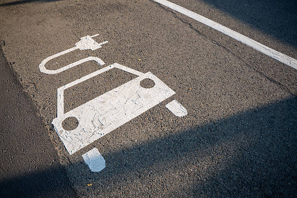 A designated parking space for electric car charging with a white painted icon on asphalt. Sunlight casts shadows on the asphalt road surface