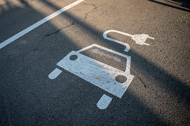 A designated parking space for electric car charging with a white painted icon on asphalt. Sunlight casts shadows on the asphalt road surface