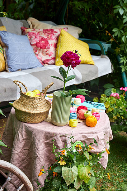 istock A small table covered with a soft pink cloth holds embroidery threads, a piece of fabric with an embroidery design, a notebook, and a glass vase with a blooming garden peony. The background features a cozy garden swing with fluffy pillows, 2220616392