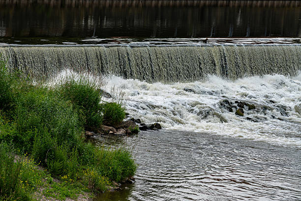 istock Waterfall cascading over a dam near a lush green riverbank 2220626836