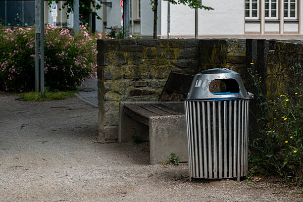 A metal trash bin sits beside a wooden bench in a serene park. The surrounding area features greenery and flowers, creating a peaceful atmosphere for visitors to enjoy.