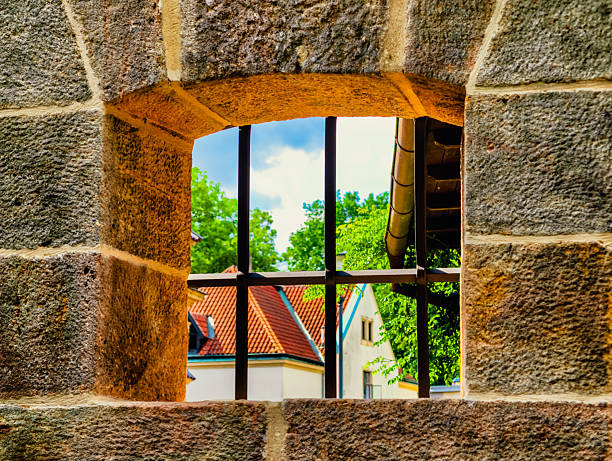 Mystical View Through an Ancient Stone Window with Iron Bars, Framing a Charming Village with Red Roofs and Lush Greenery Under a Serene Blue Sky, Perfect for Art Lovers and Decor Enthusiasts
