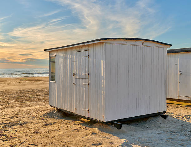 White wooden cabin on a beach, standing on soft sand, with a serene ocean and colorful sunset in the background. Captures a tranquil outdoor scene perfect for leisurely concepts.