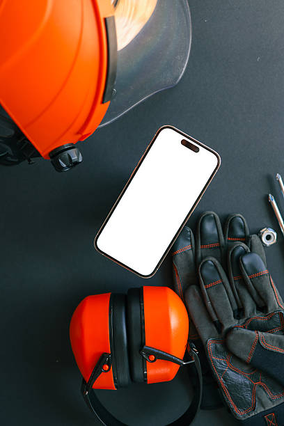 istock Smartphone with blank screen surrounded by construction tools, gloves, helmet, and ear protection on black background. Industrial safety concept 2220650359