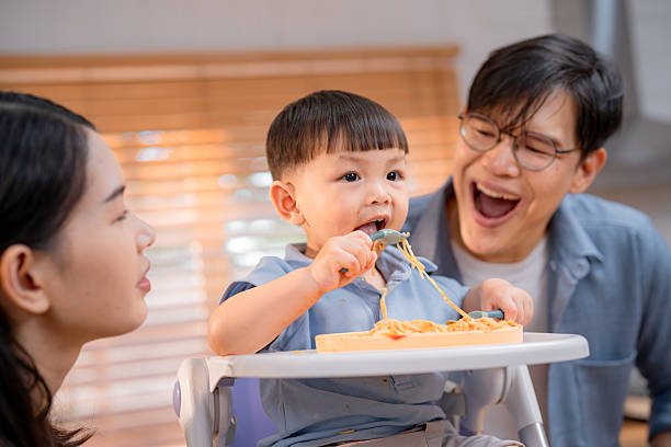 A cheerful toddler sits in a high chair, excitedly eating spaghetti. Parents look on with smiles, creating a warm and loving atmosphere in their cozy kitchen.
