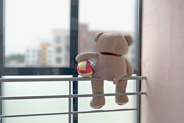 A teddy bear with a colorful ball sits perched on a balcony railing, looking out the window. The scene evokes a sense of nostalgia and adventurous imagination in a playful setting...