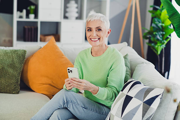 istock Senior woman using smartphone and smiling warmly while relaxing on a cozy sofa in a stylishly decorated living room 2220682632