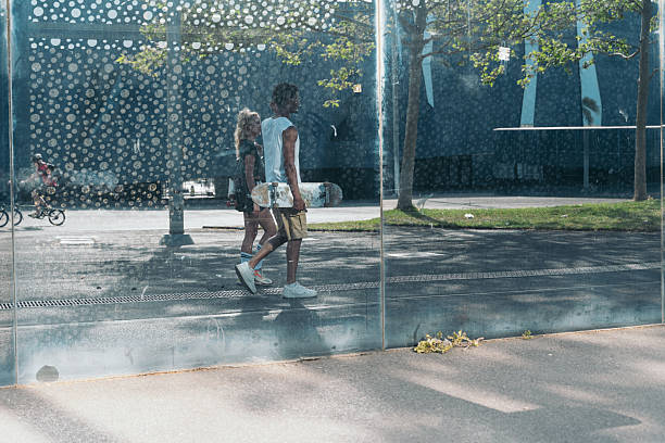 Young couple walks past a modern glass wall, the man carrying a skateboard, embodying the carefree spirit of urban youth