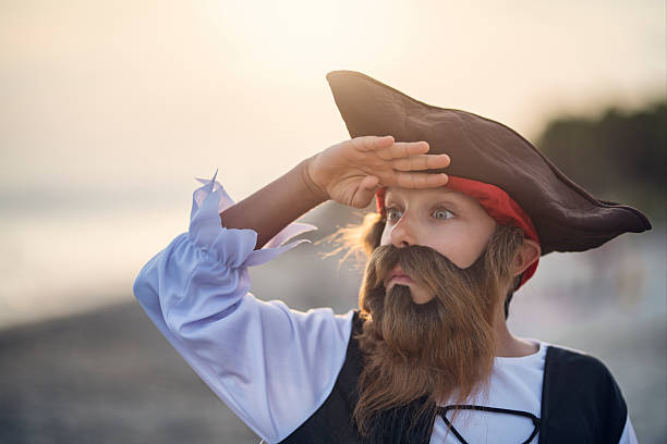 A child dressed in a classic pirate outfit stands on a beach, striking a dramatic pose while holding one hand to their forehead as if scanning the sea. The costume includes a black tricorn hat with a red bandana peeking out underneath, a white ruffled shirt, and a fitted black vest with laces. The sun casts a soft, golden glow—suggesting early morning or late afternoon—adding warmth and a touch of adventure to the seaside setting. The blurred background reveals a quiet shoreline with distant trees, allowing the young pirate to take center stage. This evocative moment channels imaginative play and storytelling, capturing the thrill of pretending to lead a high-seas expedition on a sun-drenched coast.\nShot with Nikon D850