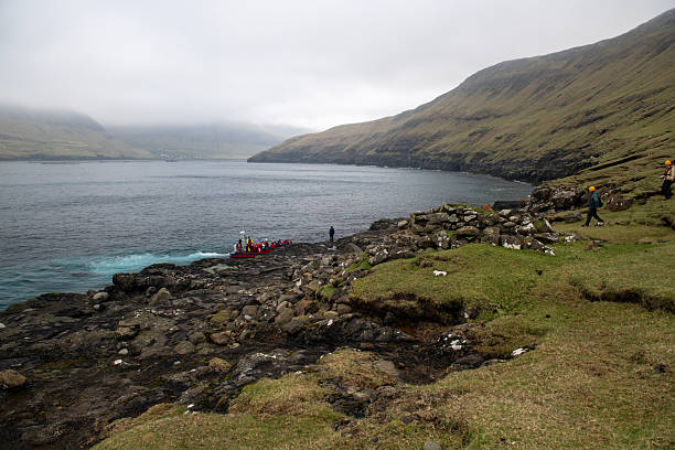 Tourists being picked up by rib boat near Drangarnir seastacks in Faroe Islands on a cloudy day. Mountains in the background.