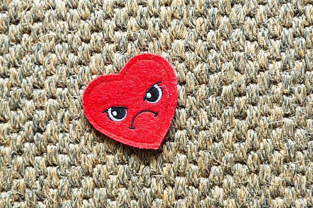 istock Red felt heart with angry expression on textured fabric. 2220693493