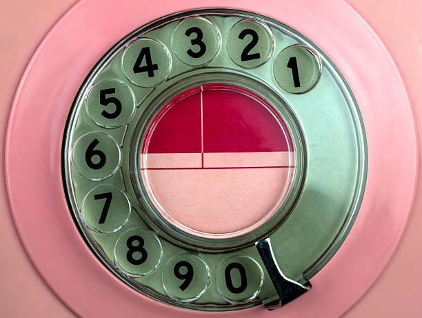 Waist up view of brunette woman in pink jumpsuit looking at camera while holding telephone and conversing with caller against matching pastel background.
