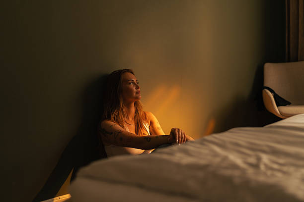 Young adult biracial woman sitting on bed with arms wrapped around knees, wearing electronic ankle monitor surrounded by food and personal items in home environment