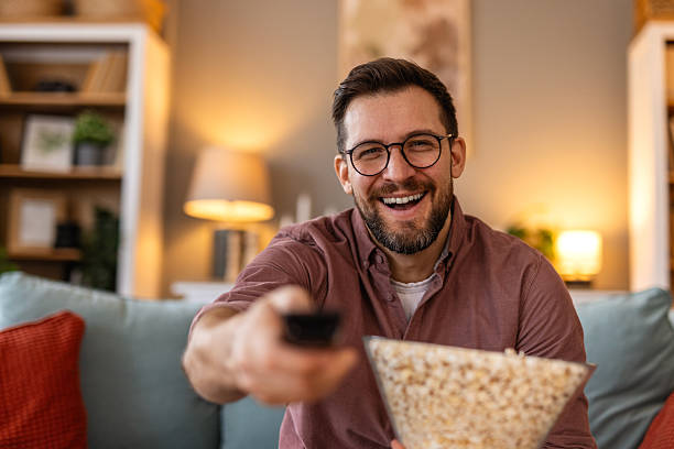 istock A joyful man sits on a comfortable couch, holding a bowl of popcorn while watching TV. His bright smile and cozy home setting create a relaxing and inviting atmosphere for a perfect night in. 2220704758