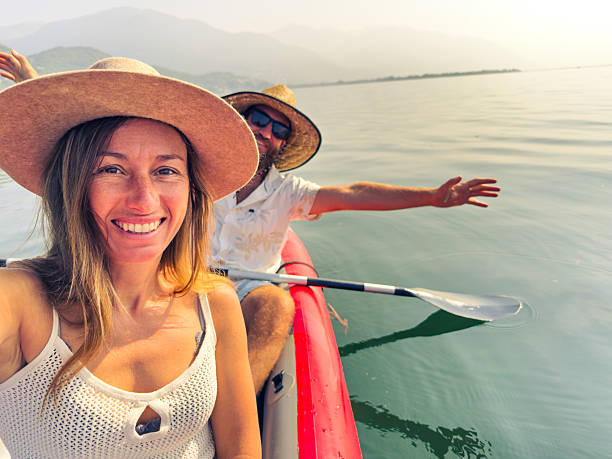 Young couple kayaking on a tranquil lake with mountains in the background. Happy woman and man in hats enjoy a sunny day in nature. Outdoor adventure and leisure activity.