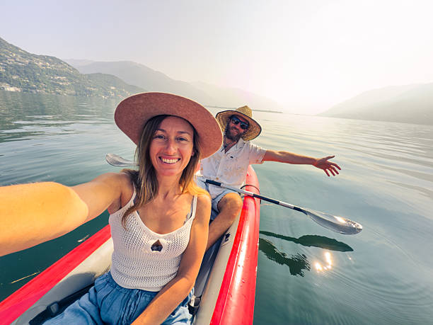 Smiling couple taking a selfie while kayaking on a serene lake. Happy pair in summer attire enjoying outdoor adventure on water. Leisurely kayaking trip amidst scenic mountain backdrop.