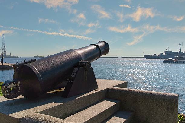 Kronstadt, Russia, September 5, 2024.An old black cannon is mounted on a stone platform overlooking a body of water. The cannon is angled towards the horizon, with a backdrop of a clear blue sky with wispy clouds. In the distance, a large naval ship is visible, with its silhouette reflected on the shimmering water. The scene is serene, with a maritime setting and historical ambiance.