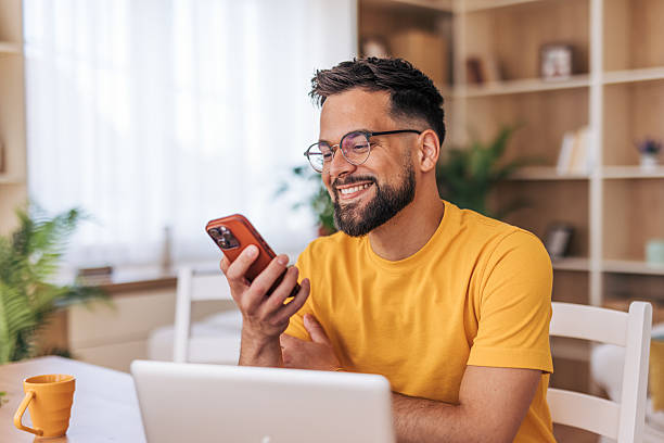 Smiling young man using smartphone and laptop at home
