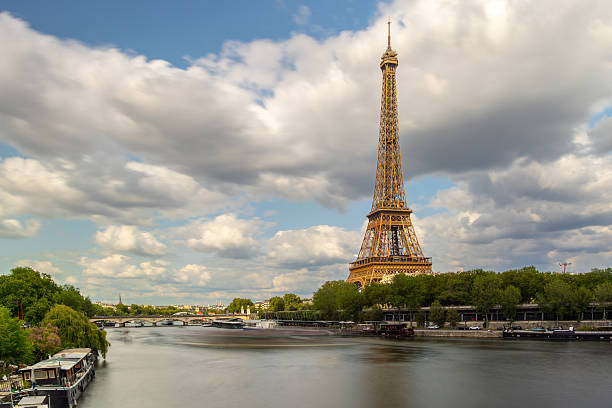 A striking long-exposure view of the Eiffel Tower rising over the Seine River in Paris, France. Boats are docked along the riverbank as dramatic clouds drift across the blue summer sky. This classic perspective captures the romance, motion, and grandeur of the French capital’s most famous landmark.