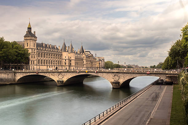 A scenic daytime view of the historic Conciergerie building and arched stone bridge over the Seine River in Paris, France. Captured under a moody, cloud-filled sky, the image showcases the Gothic architecture and cultural significance of one of Paris’s most iconic landmarks. The photo reflects European heritage, historic government institutions, and classic urban landscapes along the Île de la Cité.